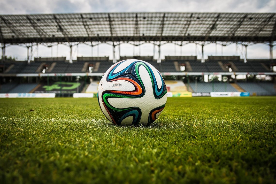 The ball at the center of the pitch — the simple object FIFA wants to keep above politics, even when the world around the stadium says otherwise. Soccer ball resting on green grass inside an empty football stadium.