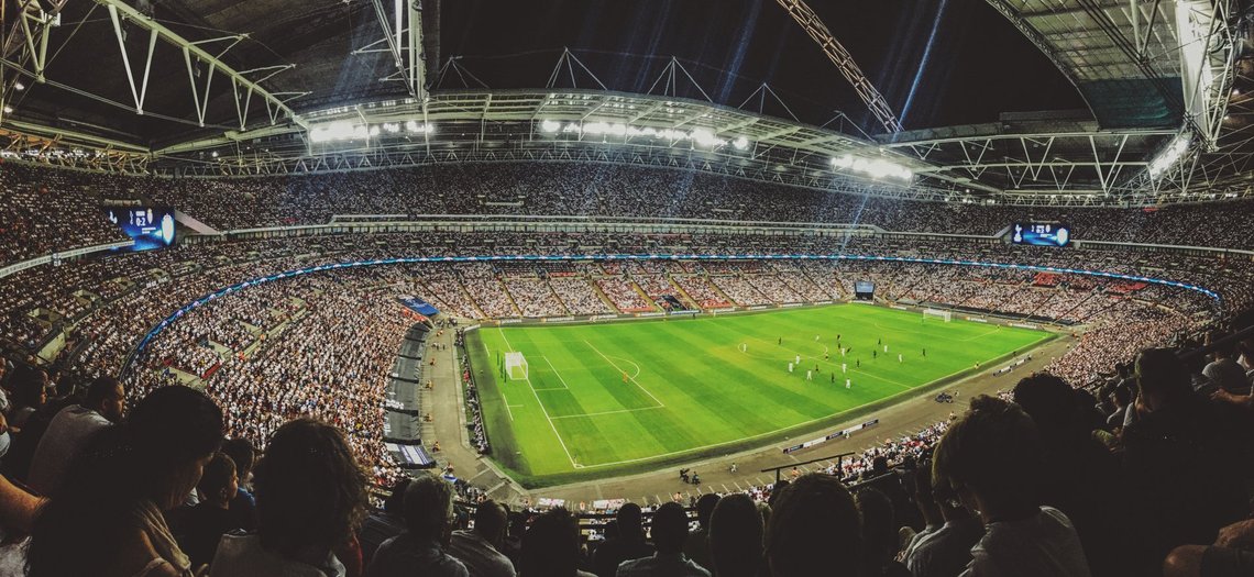 A full stadium under the lights — the stage Norway must treat as a football assignment, not just a long-awaited return. Wide-angle view of a packed football stadium at night, with floodlights, spectators filling the stands, and players on the pitch.
