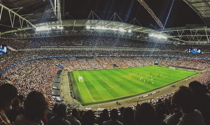 Wide-angle view of a packed football stadium at night, with floodlights, spectators filling the stands, and players on the pitch.