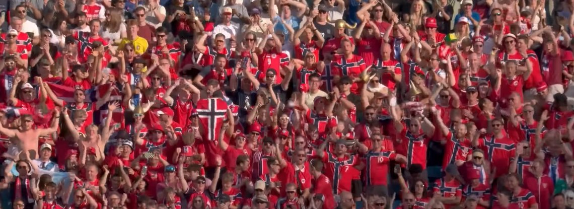 Norwegian supporters in red national team shirts cheer and wave flags from packed stadium stands during a World Cup match.