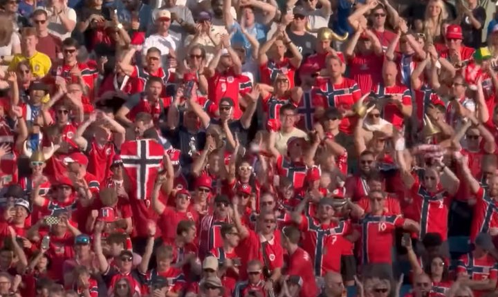 Norwegian supporters in red national team shirts cheer and wave flags from packed stadium stands during a World Cup match.