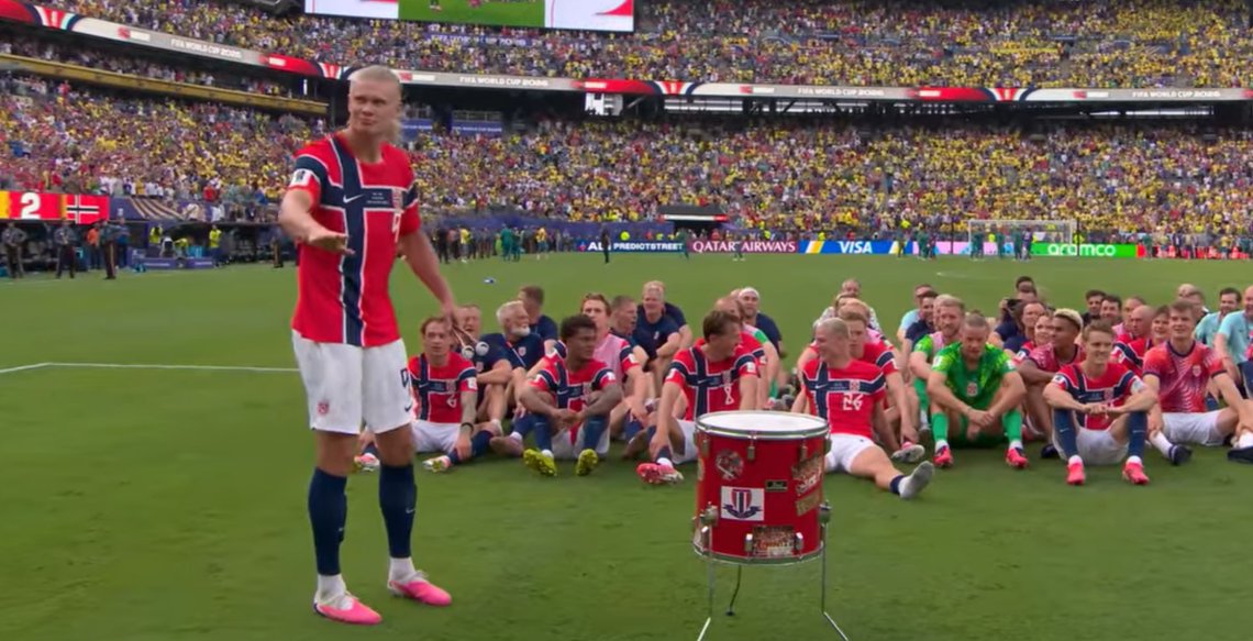 Norway’s football team sits on the pitch after beating Brazil, while one player stands beside a red drum in front of a packed stadium.