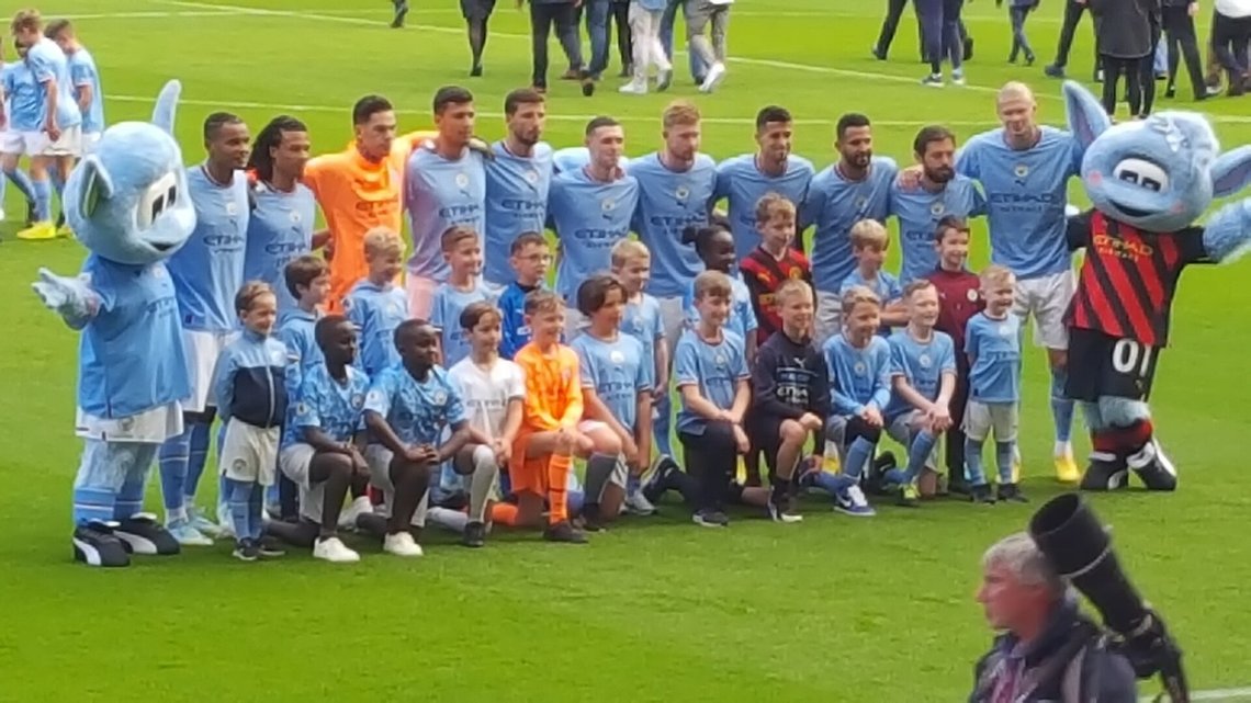 The Manchester City team, with mascots, about to face Southampton in the 2022–23 Premier League. From left to right on back row: Moonchester, Manuel Akanji, Nathan Aké, Ederson, Rodri, Rúben Dias, Phil Foden, Kevin De Bruyne, João Cancelo, Riyad Mahrez, Bernardo Silva, Erling Haaland, and Moonbeam. Photo: Wikimedia Commons The Manchester City team, with mascots, about to face Southampton in the 2022–23 Premier League. From left to right on back row: Moonchester, Manuel Akanji, Nathan Aké, Ederson, Rodri, Rúben Dias, Phil Foden, Kevin De Bruyne, João Cancelo, Riyad Mahrez, Bernardo Silva, Erling Haaland, and Moonbeam. Photo: Wikimedia Commons