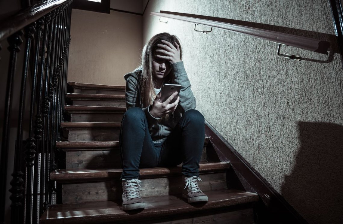 Sad depressed young teenager girl victim of cyberbullying by mobile smartphone sitting on stairs feeling lonely, unhappy, hopeless and abused. Photo: Shutterstock by SB Arts Media, Spain. Sad depressed young teenager girl victim of cyberbullying by mobile smartphone sitting on stairs feeling lonely, unhappy, hopeless and abused. Photo: Shutterstock by SB Arts Media, Spain.