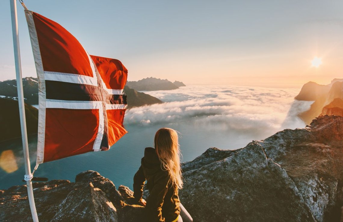 Norsk kvinne med langt blondt hår som vifter i vinden, nyter solnedgangen over fjellene og det norske flagget. Foto: Shutterstock av everst. Norsk kvinne med langt blondt hår som vifter i vinden, nyter solnedgangen over fjellene og det norske flagget. Foto: Shutterstock av everst.