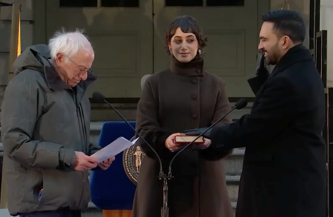 Mamdani ble innsatt som borgermester av Bernie Sanders, sammen med Mamdanis kone Rama Duwaji. Mamdani avla ed på Koranen, og ble dermed den første borgermesteren i New York City som gjorde dette. Han brukte to eksemplarer av boken: sin bestefars og Arturo Alfonso Schomburgs. Foto: Wikimedia/New York Citys borgermesterkontor. Mamdani ble innsatt som borgermester av Bernie Sanders, sammen med Mamdanis kone Rama Duwaji. Mamdani avla ed på Koranen, og ble dermed den første borgermesteren i New York City som gjorde dette. Han brukte to eksemplarer av boken: sin bestefars og Arturo Alfonso Schomburgs. Foto: Wikimedia/New York Citys borgermesterkontor.