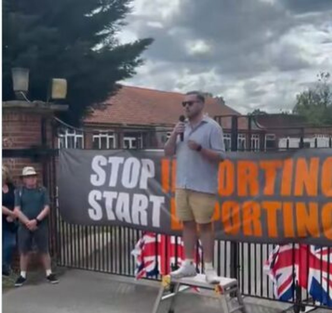 Demonstrant holder tale utenfor RAF-basen i Barnham som skal bli asylmottak. På plakaten står det «Stop importing, start deporting». Stillbilde video/ X/ Michael Hadwen Demonstrant holder tale utenfor RAF-basen i Barnham som skal bli asylmottak. På plakaten står det «Stop importing, start deporting». Stillbilde video/ X/ Michael Hadwen