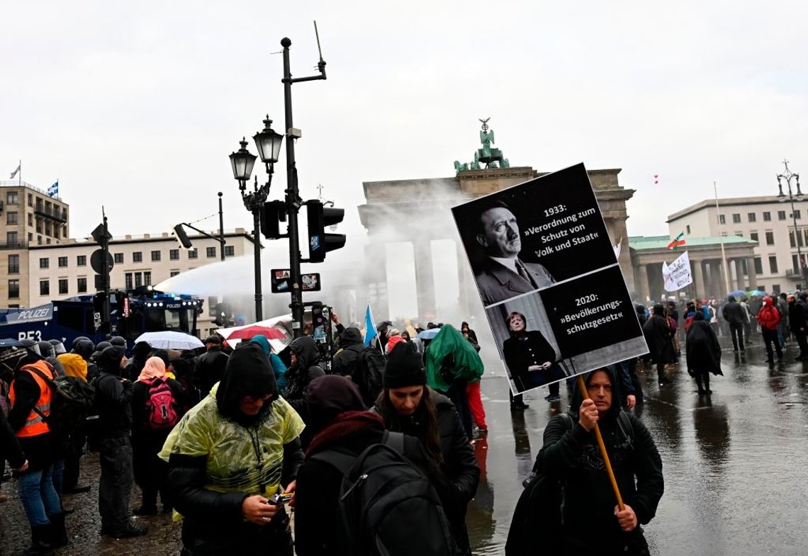 A demonstrator holds up a poster comparing Adolf Hitler's 1933 enabling act with the citizen protection law of German Chancellor Angela Merkel's government as police use a water cannon to disperse protesters demonstrating against measures imposed by the German government to limit the spread of the novel coronavirus, on November 18, 2020 at the Brandenburg Gate in Berlin. - After repeated warnings for the crowd to put on face coverings went unheeded, police said they would take action to clear the protest and "detain violators". Around 5,000 activists massed at the Brandenburg Gate, after the German government banned rallies outside parliament because of fears of violence. The demonstration mirrored similar protests seen across Europe against restrictions opponents see as a violation of their civil rights despite government warnings about the need to stop the spread of infections. (Photo by Tobias SCHWARZ / AFP) (Photo by TOBIAS SCHWARZ/AFP via Getty Images) A demonstrator holds up a poster comparing Adolf Hitler's 1933 enabling act with the citizen protection law of German Chancellor Angela Merkel's government as police use a water cannon to disperse protesters demonstrating against measures imposed by the German government to limit the spread of the novel coronavirus, on November 18, 2020 at the Brandenburg Gate in Berlin. - After repeated warnings for the crowd to put on face coverings went unheeded, police said they would take action to clear the protest and "detain violators". Around 5,000 activists massed at the Brandenburg Gate, after the German government banned rallies outside parliament because of fears of violence. The demonstration mirrored similar protests seen across Europe against restrictions opponents see as a violation of their civil rights despite government warnings about the need to stop the spread of infections. (Photo by Tobias SCHWARZ / AFP) (Photo by TOBIAS SCHWARZ/AFP via Getty Images)