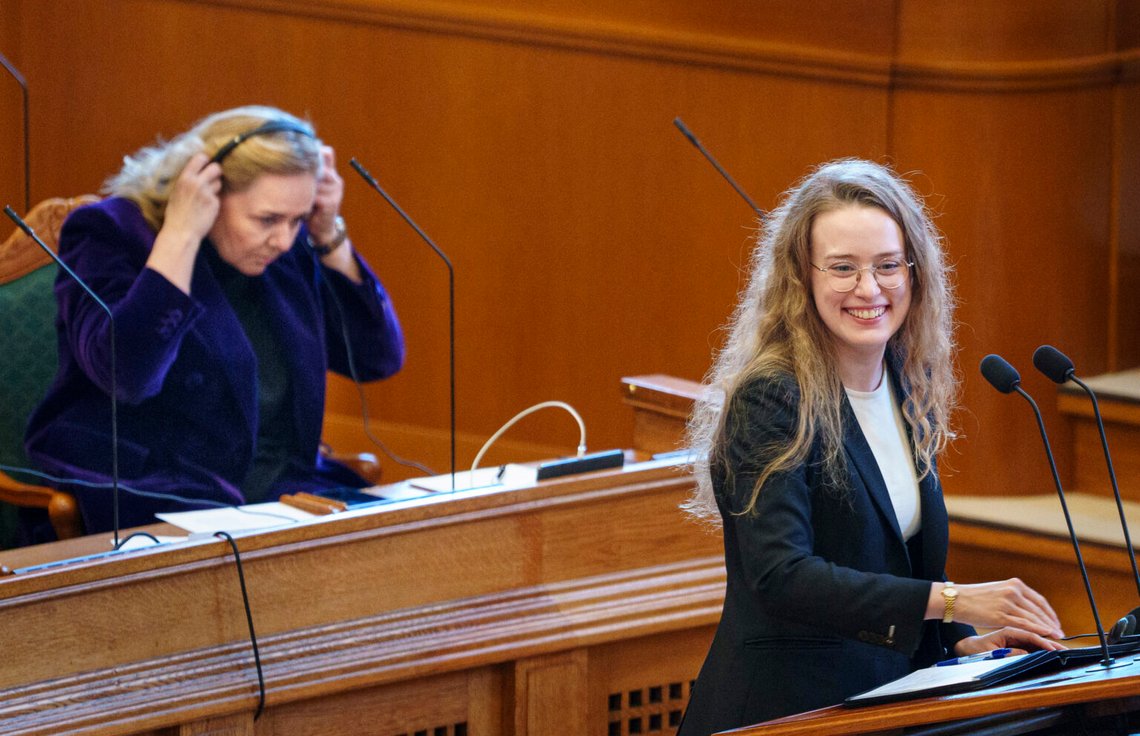 Statsminister Mette Frederiksen (S) och Siumut Aki-Matilda High-Dam i bakgrunden under förlängd frågestund i danska riksdagssalen i Christiansborg, Köpenhamn, 26 november, Foto: Liselotte Sabroe / Ritzau Scanpix Statsminister Mette Frederiksen (S) och Siumut Aki-Matilda High-Dam i bakgrunden under förlängd frågestund i danska riksdagssalen i Christiansborg, Köpenhamn, 26 november, Foto: Liselotte Sabroe / Ritzau Scanpix